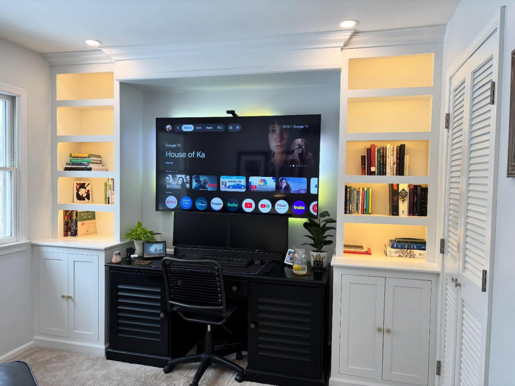 White built-in cabinets and illuminated open shelves framing a desk and television in a home office